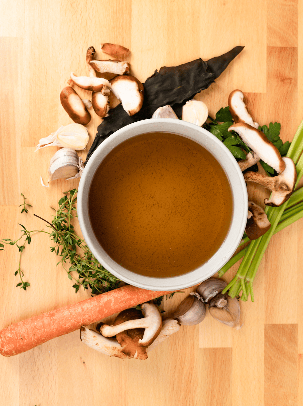 Cup of broth surrounded by vegetables on a wooden surface