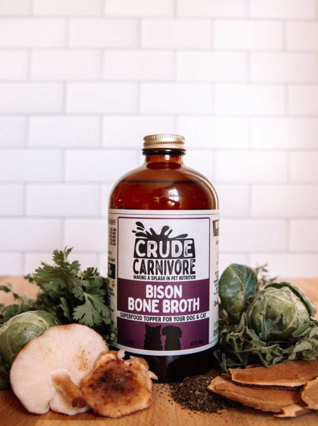Bottle of Crude Carnivore Bison Bone Broth on a wooden surface with herbs and vegetables.