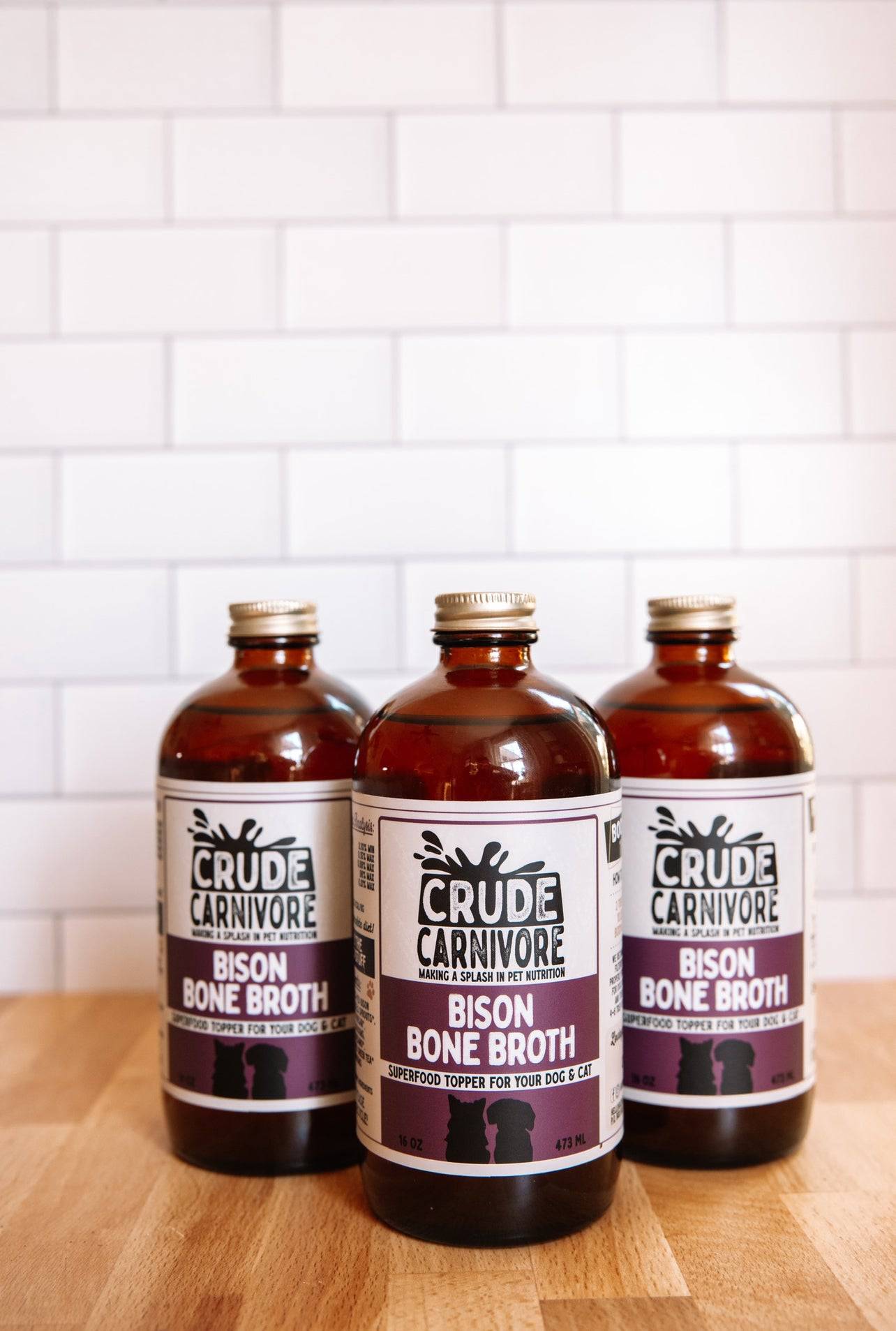 Three bottles of Crude Carnivore Bison Bone Broth on a wooden surface with a white tiled wall background.
