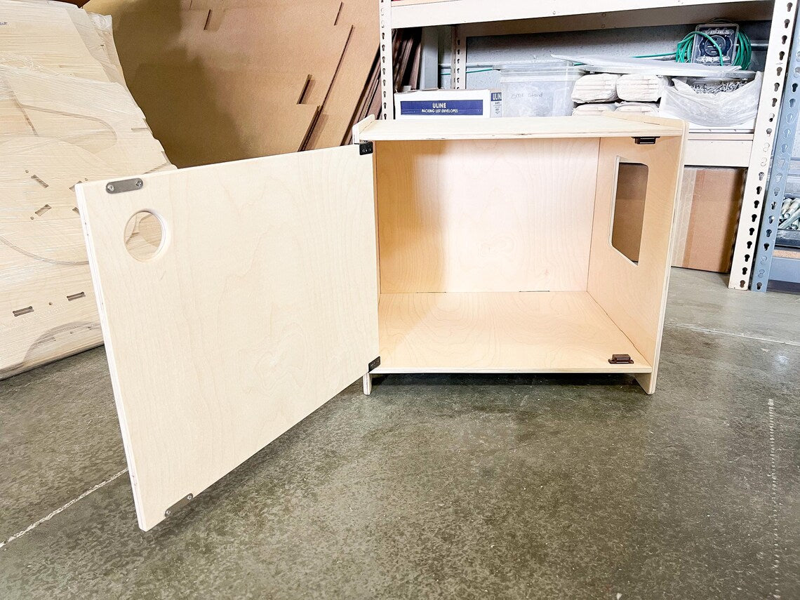 Wooden cat litter box storage unit with a door on a concrete floor, possibly in a warehouse setting.