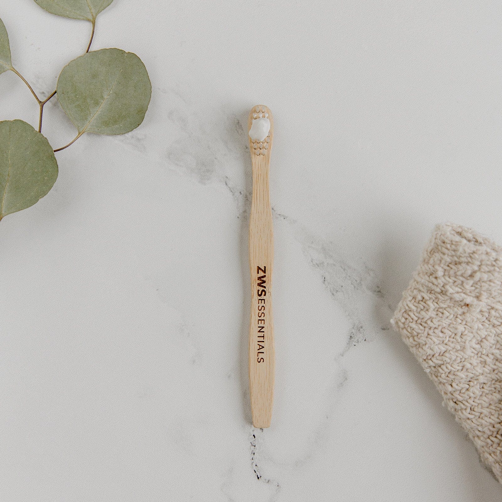 Bamboo toothbrush with 'ZWS Essentials' branding on a marble surface with a sponge and eucalyptus leaves.