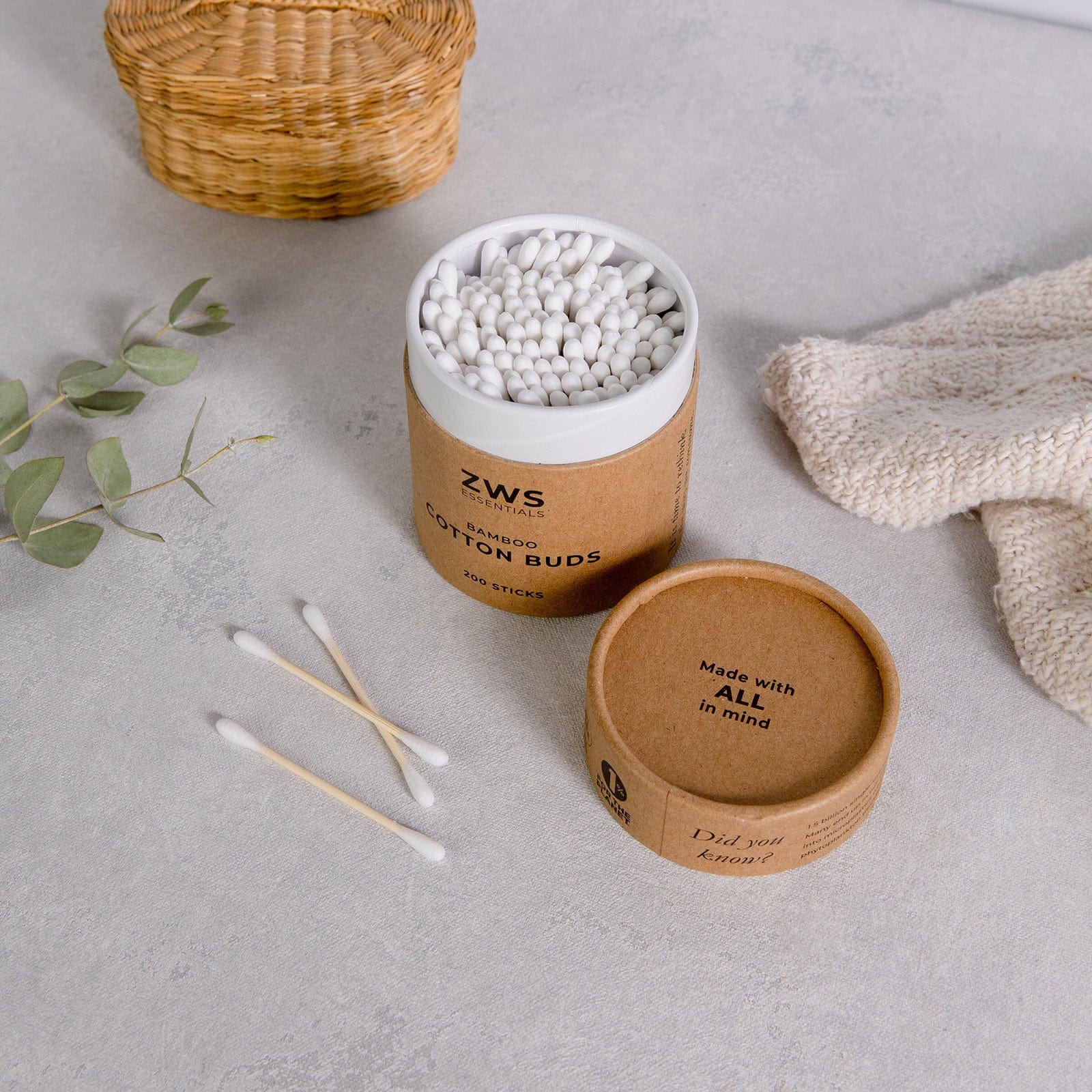 ZWS cotton buds container with cotton buds on a light gray surface with a wicker basket and greenery.