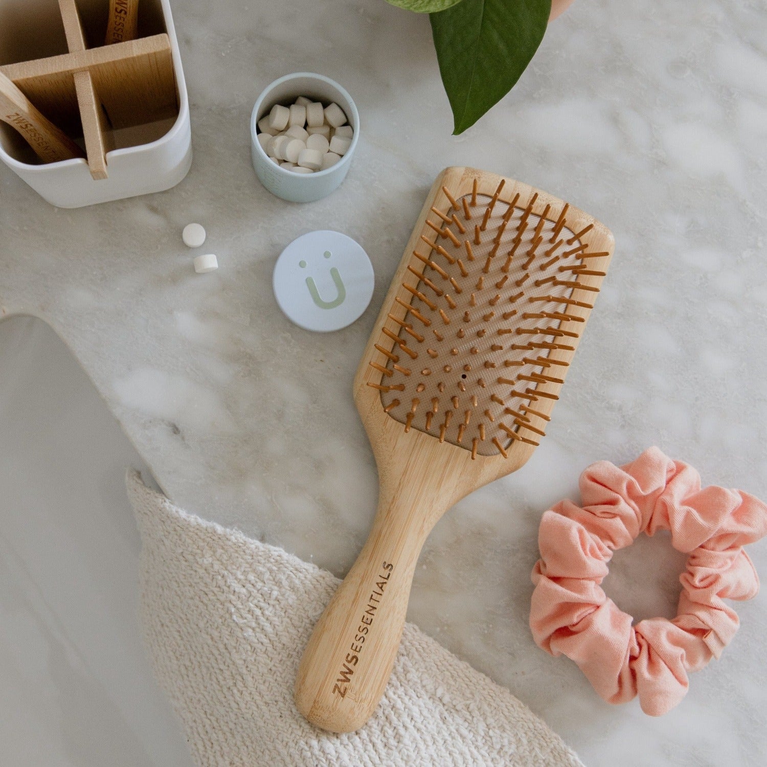 Wooden hairbrush and pink scrunchie on a marble surface with a container and small bowl in the background.