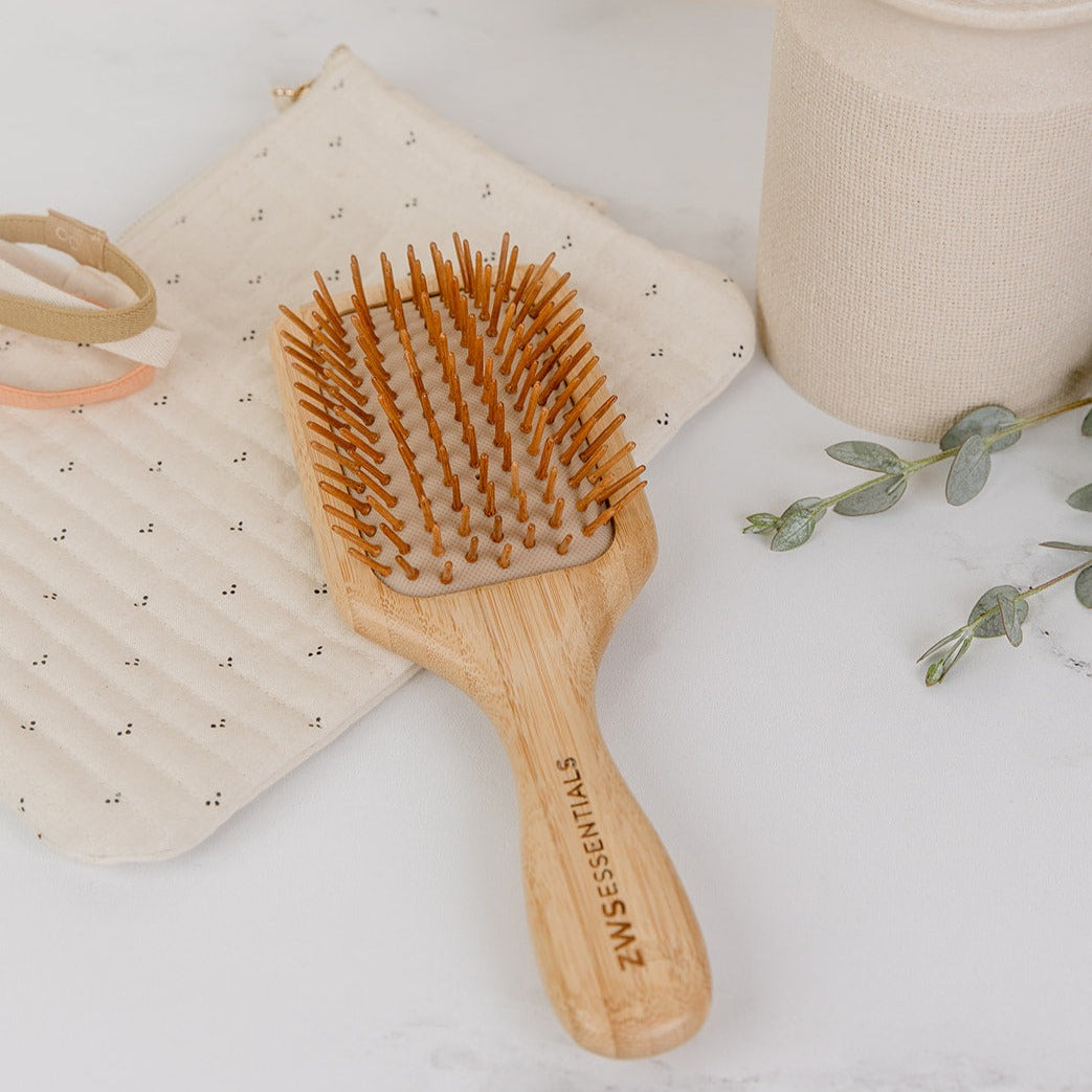 Wooden hairbrush with bristles on a white surface, surrounded by a beige pouch and green leaves.